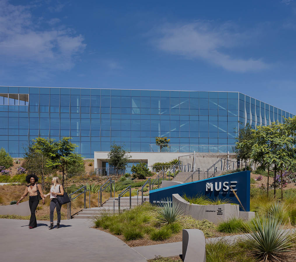 The entrance to the Muse at Torrey Pines property at 3030 Science Park in San Diego, California with the Muse sign, native landscaping, and two glass covered buildings. The buildings are owned by DHC and are LEED Silver Certified