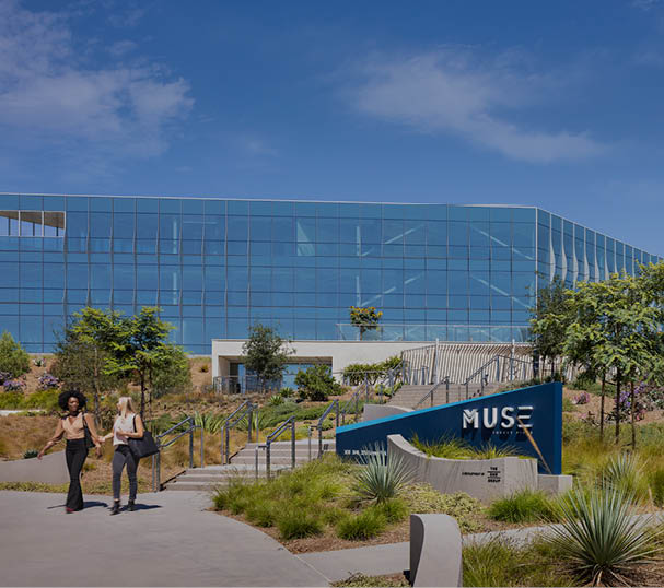 The entrance to the Muse at Torrey Pines property at 3030 Science Park in San Diego, California with the Muse sign, native landscaping, and two glass covered buildings. The buildings are owned by DHC and are LEED Silver Certified